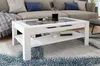 A white coffee table with a glass insert is shown in a living room setting, with a sofa in the background. Produktbild-Vorschau