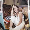 A young woman sits in a vehicle filled with stuffed animals, wearing white overalls and pink earbuds. Produktbild-Vorschau
