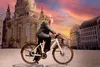 A woman rides a white e-bike in a European city square with a large church in the background and a colorful sunset sky. Produktbild-Vorschau