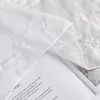 A close-up view of white embroidered sheer fabric, partially covering a book with text. Produktbild-Vorschau