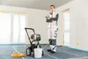 A painter stands in a room under construction, holding a paint sprayer. A paint sprayer machine and a bucket are on the floor. Produktbild-Vorschau