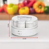 A white yogurt maker is shown on a white countertop, with a blurred background of colorful fruits and vegetables. Produktbild-Vorschau