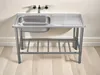 A stainless steel sink with a drainboard and a metal stand, set against a light-colored wall and wooden floor. Produktbild-Vorschau