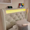 A close-up view of a beige upholstered bed with integrated LED lighting, a smartphone charging, and a tablet on the headboard. Náhled obrázku produktu