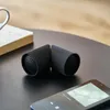 Two black cylindrical speakers are placed on a dark table next to a smartphone and a book. Podgląd zdjęcia produktu