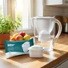 A water filter pitcher and its replacement cartridges are displayed on a kitchen countertop next to a bowl of fruit and a potted plant. Produktbild-Vorschau
