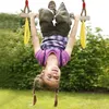 A young girl is hanging upside down from a wooden trapeze swing with yellow plastic rings, outdoors in a grassy area with trees in the background. Produktbild-Vorschau
