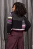 A woman wearing a colorful knitted sweater and dark pants stands with her back to the camera. Produktbild-Vorschau