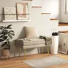 A beige upholstered bench with gold legs sits in a room with a textured white wall and wooden stairs. Anteprima dell’immagine prodotto