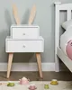 A white nightstand with wooden legs and bunny ears, next to a bed. A toy tea set is on the floor in front of it. Náhľad obrázku produktu
