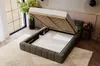 A dark gray upholstered bed with its storage compartment open, revealing the slatted base and storage space. The bed is placed on a patterned rug in a well-lit room with large windows and plants. Produktbild-Vorschau
