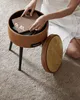 A person's hands are placing a book into a round brown storage ottoman with wooden lid off to the side, resting on a plush carpet. Náhled obrázku produktu