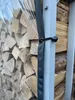 A close-up view of a transparent PVC cover for a firewood shelter, secured with a black strap and toggle to a gray metal pole. Piles of firewood are visible behind the cover. Produktbild-Vorschau