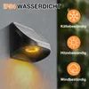 A solar-powered outdoor wall light is shown mounted on a gray wall during a rain shower. The light emits a warm yellow glow. To the right, there are icons and text indicating resistance to cold, heat, and wind. Produktbild-Vorschau
