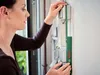 A woman is using a Bosch measuring tool to measure the height of a light switch on a wall. Produktbild-Vorschau