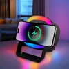A smartphone is wirelessly charging on a black stand that also functions as a Bluetooth speaker and has colorful LED lighting. The background shows a dimly lit room with furniture and a window. Produktbild-Vorschau