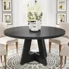 A round black dining table with a unique cross-leg base is set in a dining room with chairs, artwork on the wall, and a large window with sheer curtains. Anteprima dell’immagine prodotto