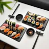 A sushi serving set with two black slate platters, sushi, chopsticks, and soy sauce dishes, arranged on a white surface. Voir les images du produit