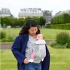 A woman is carrying a baby in a carrier in a park. Anteprima dell’immagine prodotto