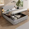 A bedroom scene featuring a hydraulic storage bed with its storage compartment open, revealing stored items. The bed is made up with bedding and pillows, and a plant is visible in the background. Produktbild-Vorschau