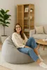 A woman is sitting on a large, light gray beanbag chair in a living room. She is smiling and looking at the camera. The room has a wooden bookshelf, a sofa, and a plant. Produktbild-Vorschau