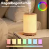 A humidifier diffuser with colored lights is shown on a nightstand next to a bed, with smaller versions of the diffuser displaying different colors in the foreground. Produktbild-Vorschau