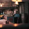 Two people are sitting at a wooden bar in a rustic-themed room, with a fireplace and a barrel visible. Produktbild-Vorschau