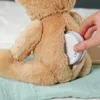 A hand is inserting a white circular device into a slit on the back of a beige teddy bear. Anteprima dell’immagine prodotto