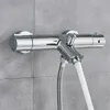 A chrome thermostatic shower faucet with a white shower hose attached, mounted on a gray tiled wall. Náhľad obrázku produktu