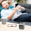 A man is lying on a couch, looking at his phone, with a small speaker and glasses on a table in the foreground. Musical notes are depicted floating from the speaker. Produktbild-Vorschau