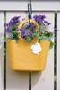 A yellow hanging planter filled with purple flowers and green leaves, attached to a white wooden fence. Voir les images du produit