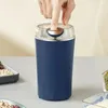 Dunkelblauer elektrischer Mühle mit durchsichtigem Deckel, eine Hand drückt den goldenen Knopf oben, daneben Schalen mit Bohnen Náhľad obrázku produktu