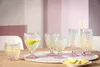 A collection of glasses with colorful bases and textured bodies, some containing drinks and garnishes, arranged on a table. Produktbild-Vorschau