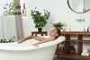 A woman is relaxing in a bathtub in a well-lit bathroom, with plants and toiletries visible in the background. Produktbild-Vorschau