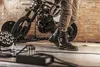 A person wearing motorcycle boots stands next to a motorcycle in a studio setting with brick walls and lighting equipment. Podgląd zdjęcia produktu