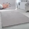 A hand is shown pressing into a soft, gray, high-pile rug, demonstrating its plushness. The rug is laid out on a light wooden floor in a room with modern furniture. Produktbild-Vorschau