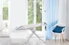 An ironing board is set up in a bright room with large windows and sheer blue curtains. Náhľad obrázku produktu