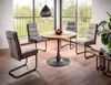 A dining set with a round wooden table and four chairs in a room with a large window overlooking greenery. Voir les images du produit