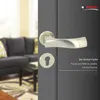 A door handle and lock plate are shown on a gray door, with a blurred living room in the background. Produktbild-Vorschau