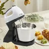 A stand mixer with a stainless steel bowl is shown on a table with baked goods and flowers in the background. Produktbild-Vorschau