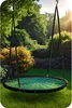 A round swing seat with a blue net pattern hangs in a lush garden setting with trees and flowers. Podgląd zdjęcia produktu