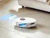 A white robot vacuum cleaner on a tiled floor next to a bed. Voir les images du produit