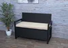 A black poly-rattan storage bench with a beige cushion sits on a wooden floor in front of a wood-planked wall. A potted plant is to the left of the bench. Voir les images du produit