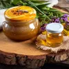 Two jars of honey are displayed on a wooden surface, surrounded by greenery and small purple and yellow flowers. Podgląd zdjęcia produktu