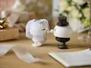 Wedding-themed decorative figures on a wooden surface with gifts and a notebook in the background. Produktbild-Vorschau