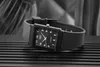A black Casio watch with a rectangular face and a black strap is placed on a black ribbed surface. The background is blurred and consists of gray and black tones. Anteprima dell’immagine prodotto