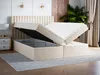 A bed with its storage compartment open, revealing the interior space. The bed is upholstered in a beige fabric and has a tall, fluted headboard. A bedside table with drawers and a lamp is visible to the left, and curtains and a plant are in the background Produktbild-Vorschau