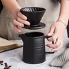 Hands holding a black ceramic pour-over coffee maker and filter set over a matching carafe. Produktbild-Vorschau