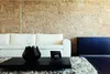 A modern living room with a white sofa, a black coffee table with a vase and a small electronic device, and a dark cabinet against a stone wall. Anteprima dell’immagine prodotto