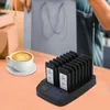 A coffee cup sits on a table next to a wireless pager system with multiple pagers in a charging base. In the background, a person is holding shopping bags. Voir les images du produit